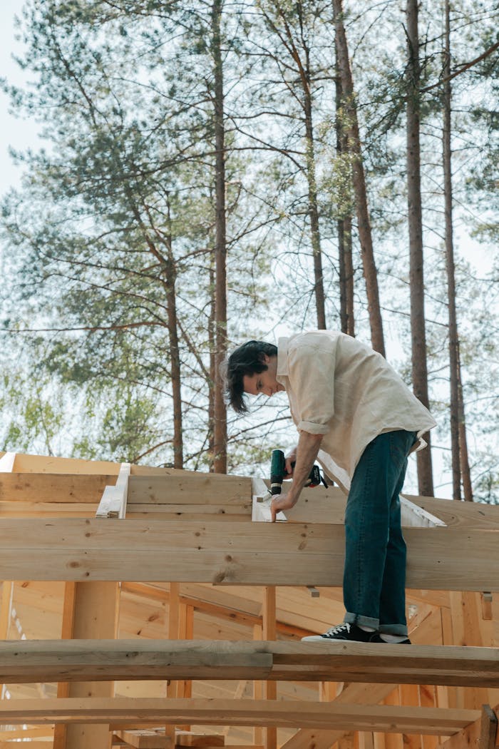 Man working on wooden house construction amidst tall trees, drilling on roof.