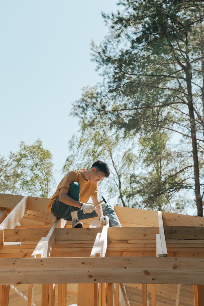 A carpenter works on a wooden roof frame outdoors under clear skies.